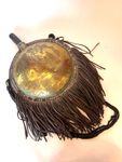 GOURDE de forme demi-sphérique, la panse en laiton à décor incisé d'un dromadaire dans un paysage orientaliste. Le contour clouté, orné de franges en cuir. Le goulot en bois et plaque de cuivre. L'ans