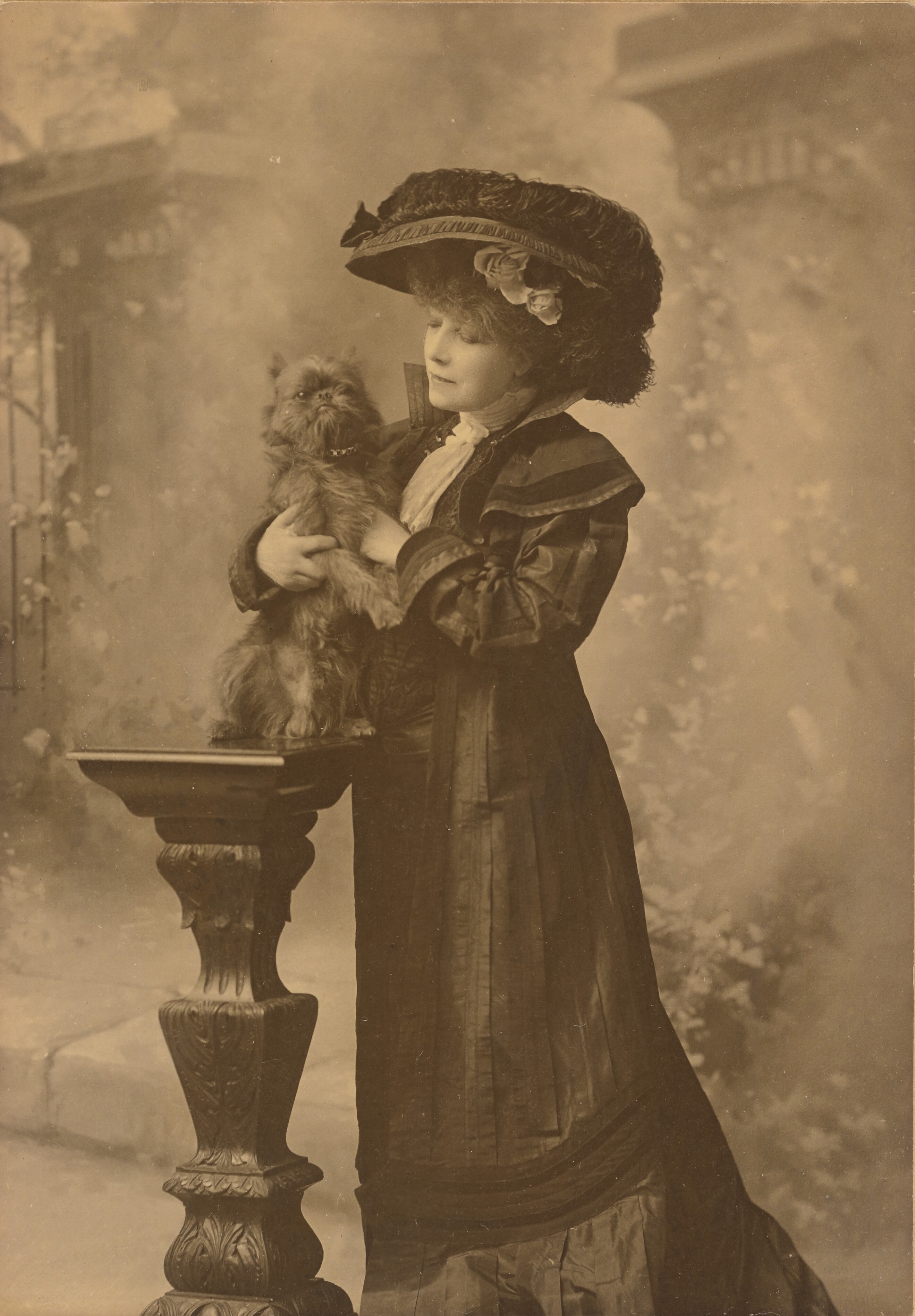 [Sarah Bernhardt standing in a studio setting, holding a little black dog on an ornate, carved wooden pedestal]