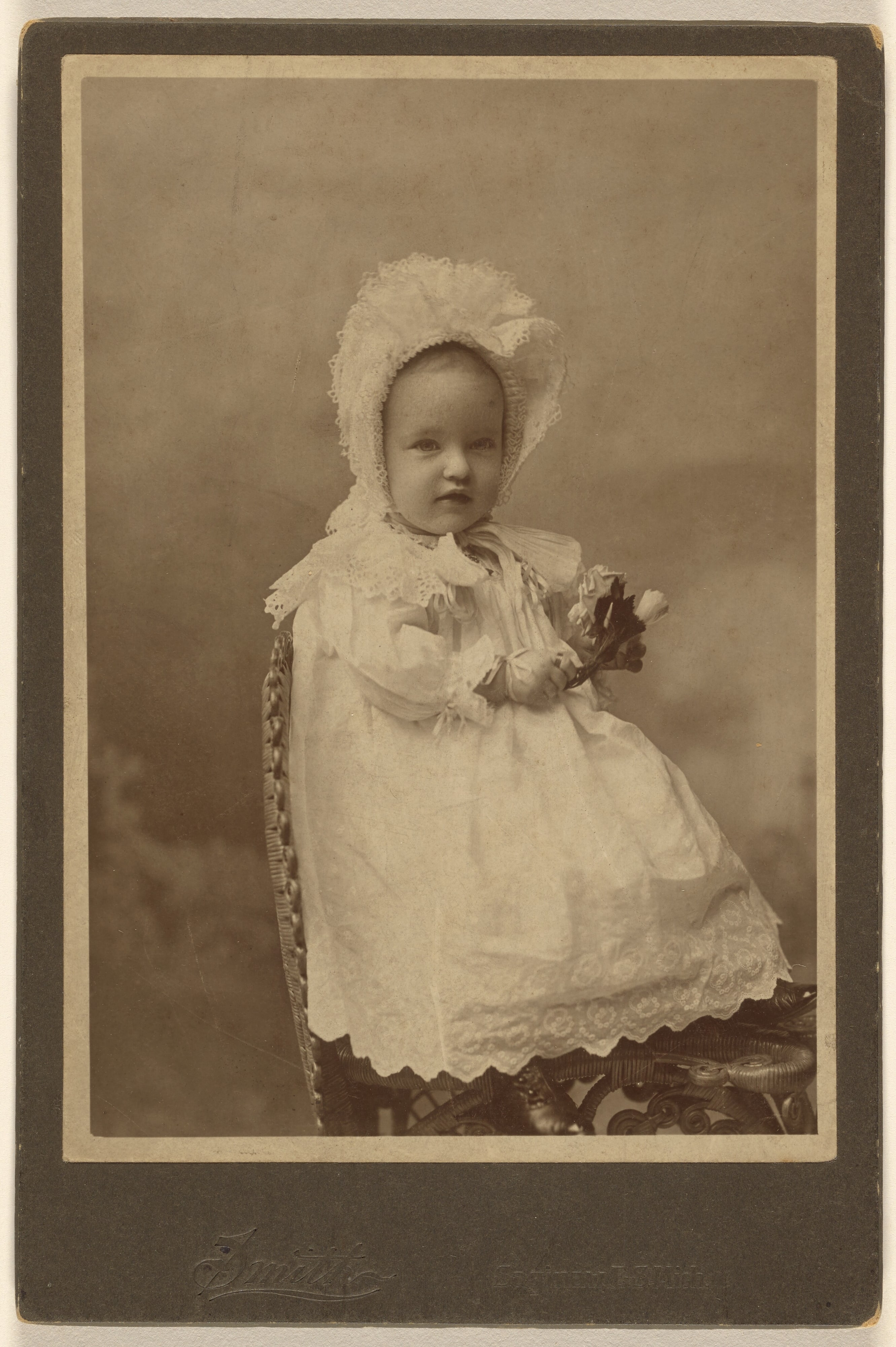 [Unidentified baby wearing a bonnet, holding a flower, seated]