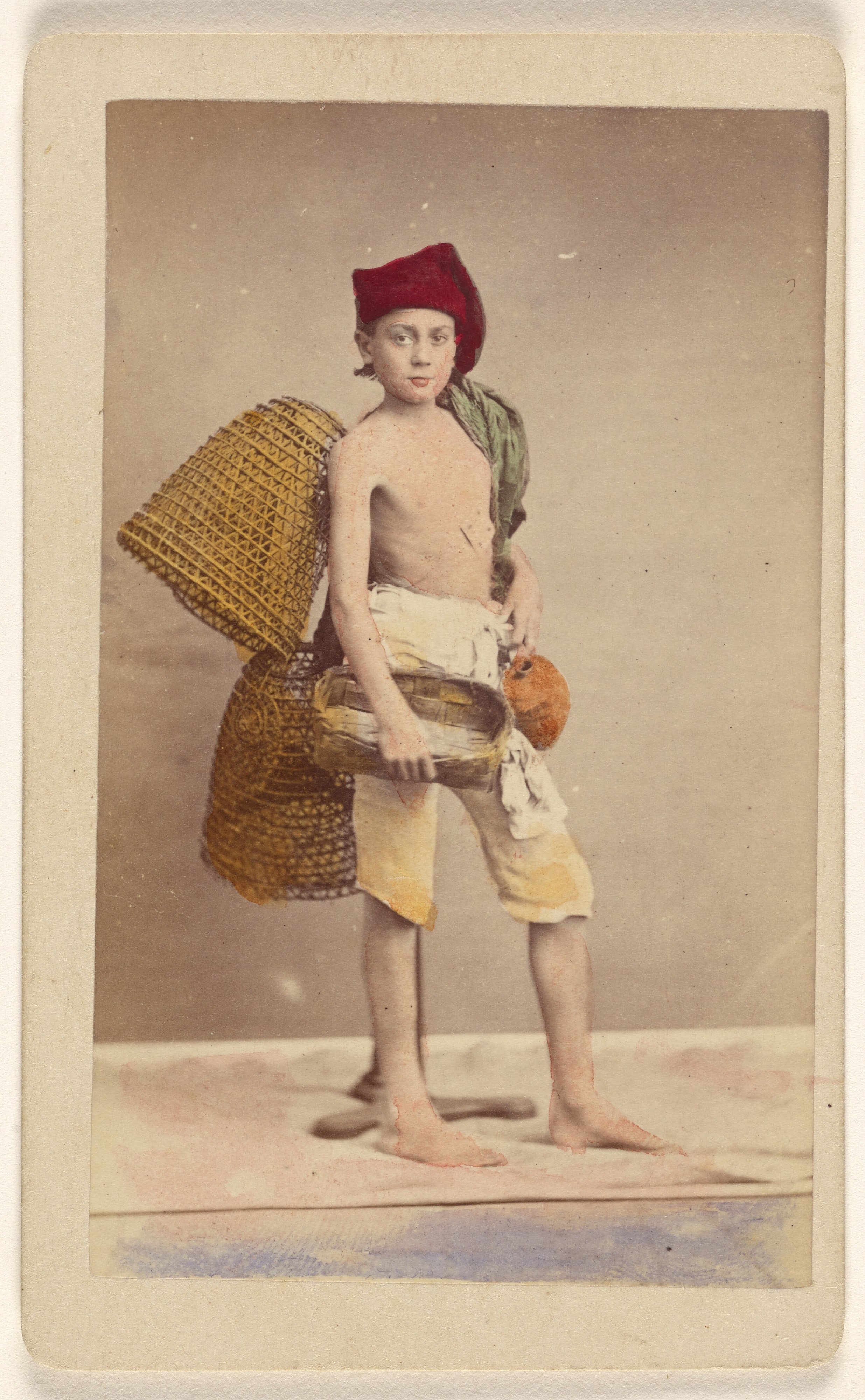 [Unidentified boy wearing native costume with baskets on back and carrying a water jug and small basket, standing]