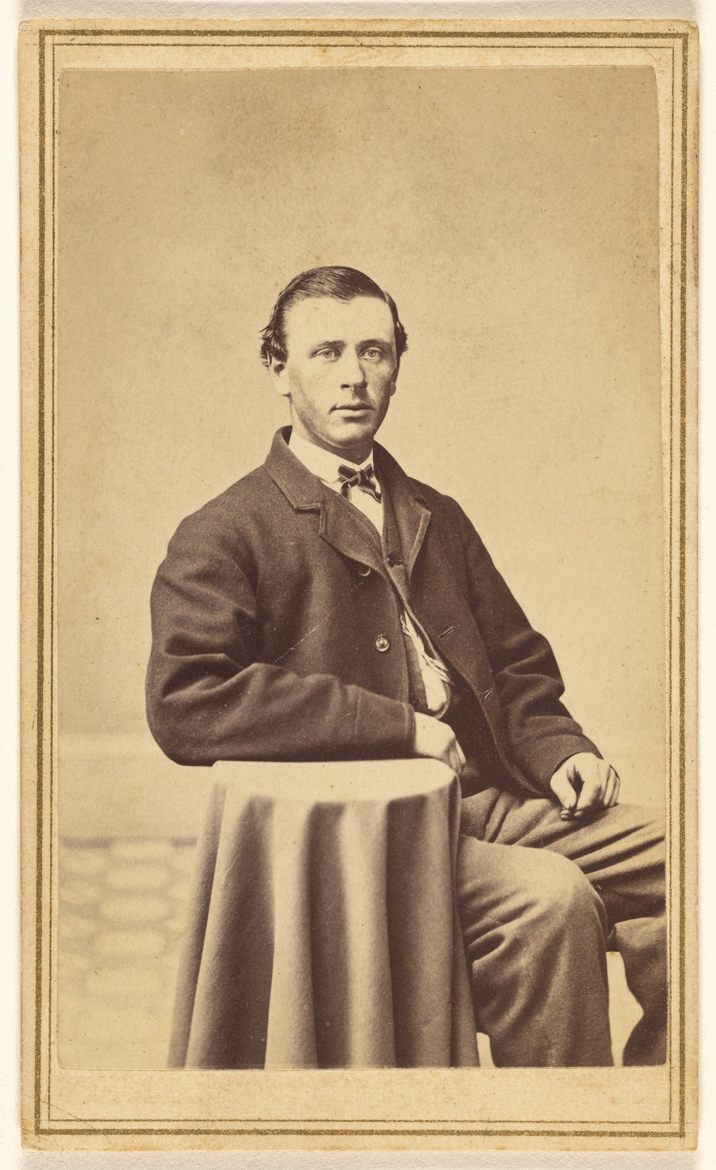 [Unidentified man seated, with arm on small covered table]