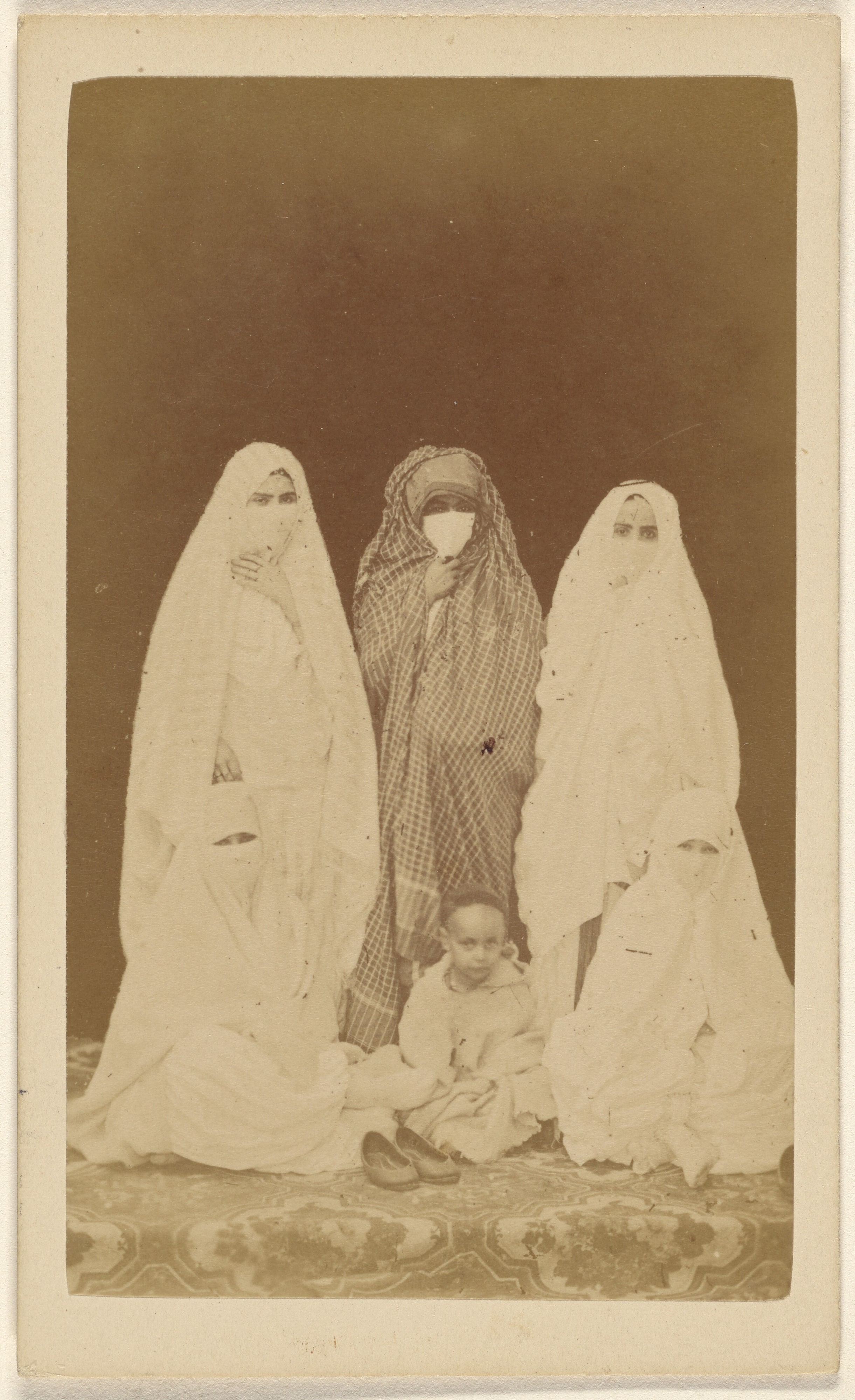 [Three Algerian women wearing veils standing, with a little unveiled girl, seated at their feet]