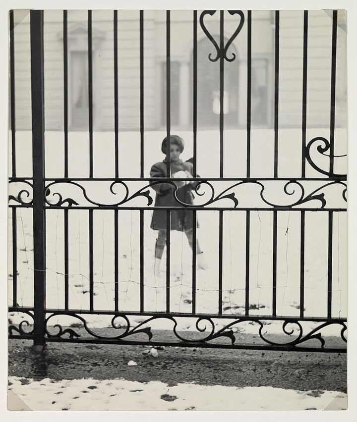 “Al di La” (girl making snowball behind iron fence)