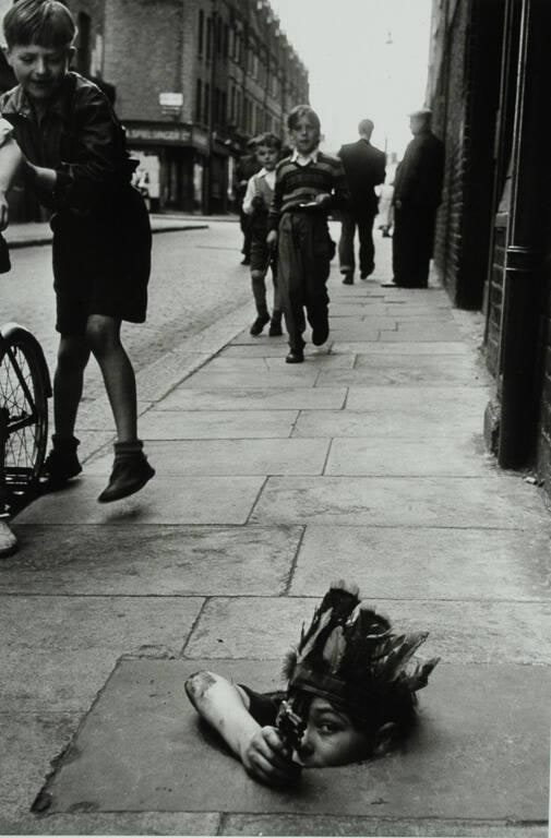 A young child wearing an Indian headdress hides in a coal hole as he takes aim under the watchful eye of a friend.