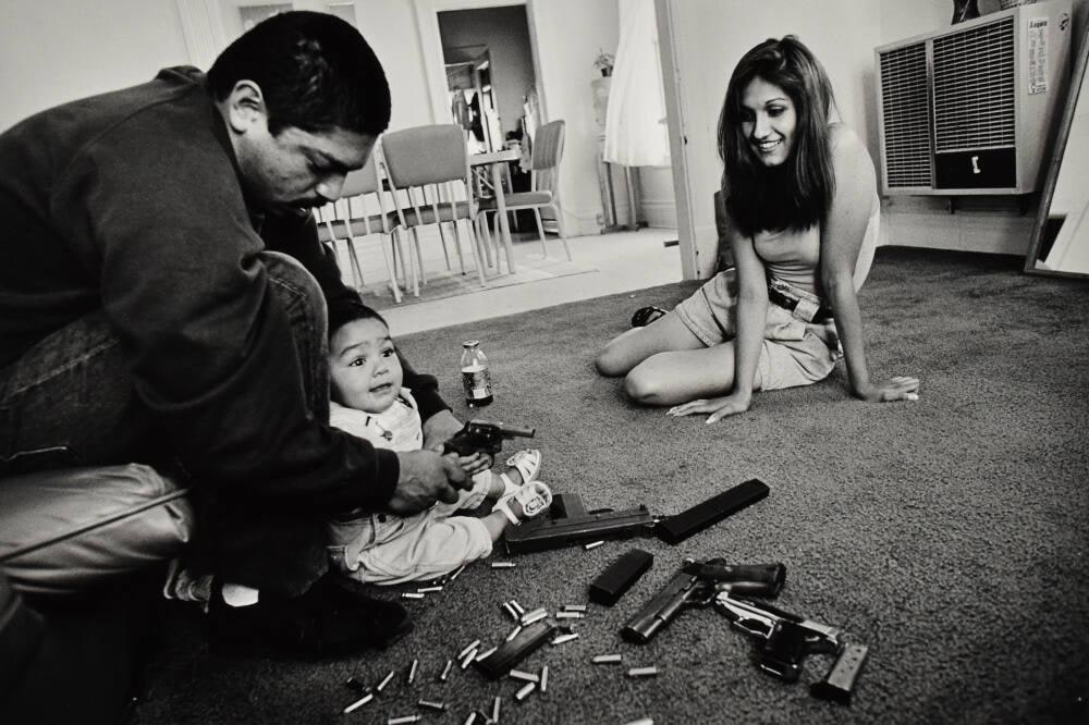 The morning after a rival gang tried to shoot Chivo for the fourth time. Chivo teaches his daughter how to hold a .32-caliber pistol. Her mother, Yvonne, looks on. Boyle Heights, Los Angeles, CA