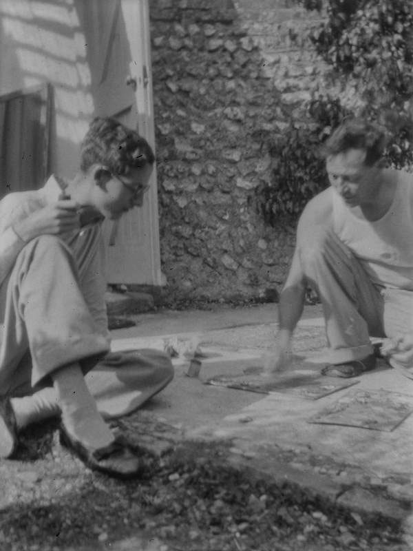 ‘Black and white negative of Eddy Playfair and Duncan Grant kneeling down, looking at some paintings on the ground in the garden at Charleston, Firle, Sussex