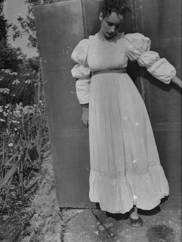 ‘Black and white negative of Angelica Bell wearing a white Victorian dress, standing in front of a screen in the garden at Charleston, Firle, Sussex