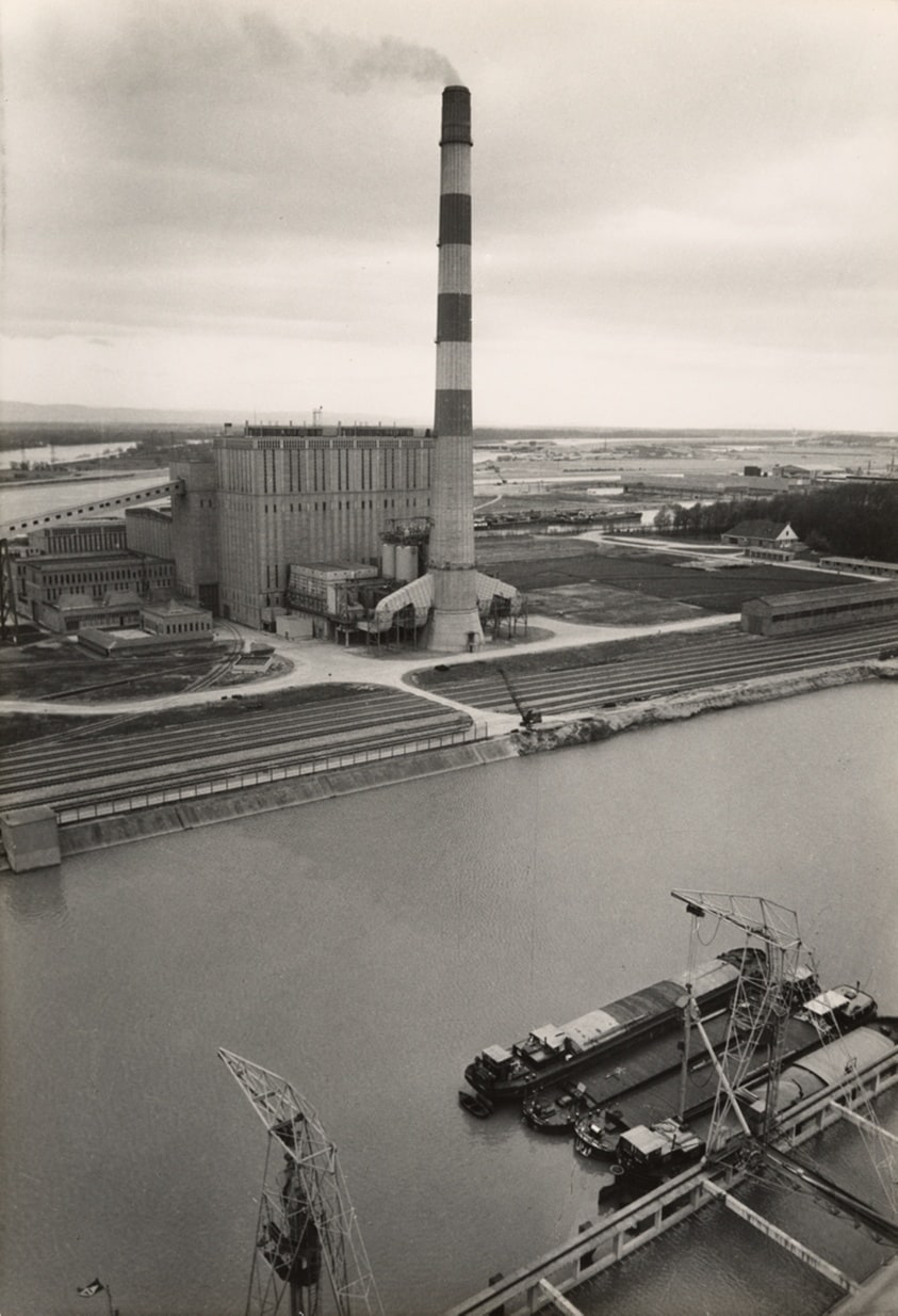 The developments of the European economy have required Strasbourg to maintain its transit function, modernize the machines in its warehouses, increase the surface of its roads, and open new docks. These are placed south of the city at the mouth of the Grand Canal d'Alsace. From the top of the large silo, which was built a few years ago, one can see the powerful Central Thermonique N°2, recently built by Electricity of France